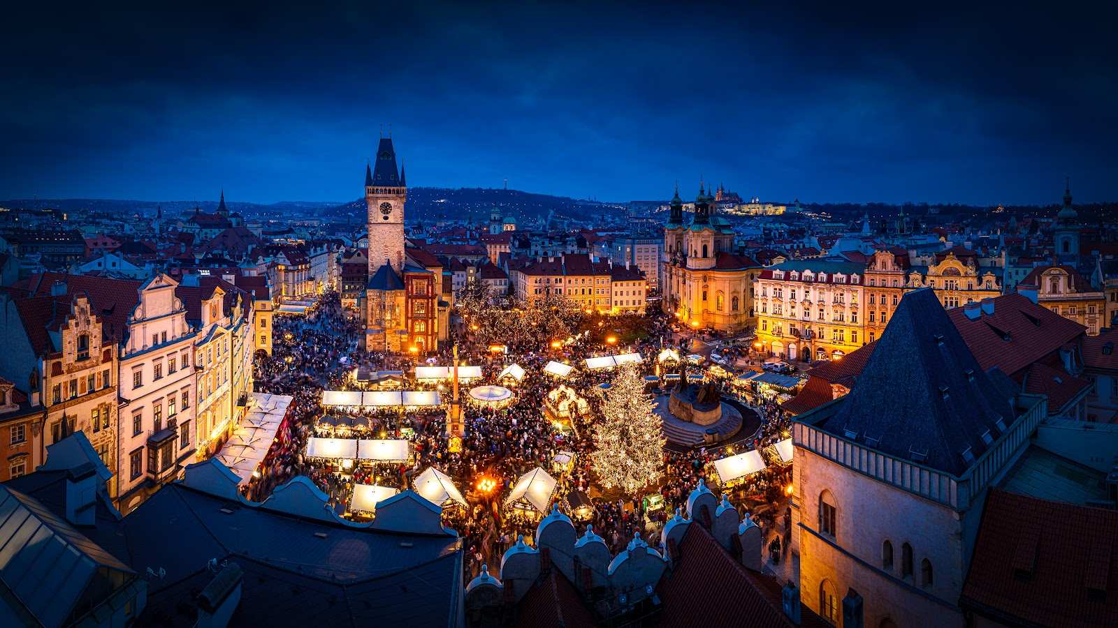 Der Weihnachtsbaum und der Markt auf dem Altstädter Ring in der Dämmerung, dahinter der Altstädter Rathausturm und die Prager Burg.